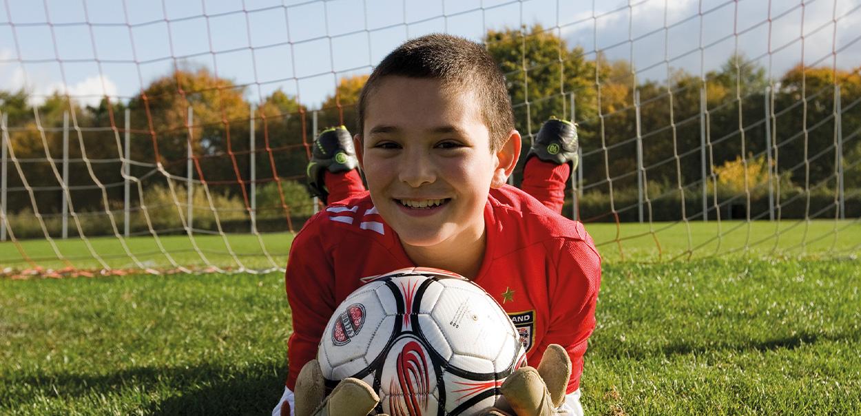 Dreng ligger på maven med bold i hænderne foran fodboldmål