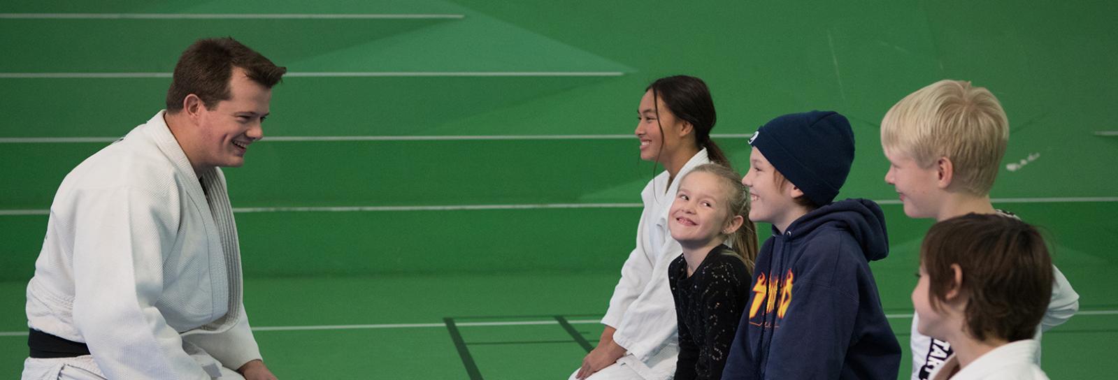 Børn sidder overfor taekwondo træner i træningssal